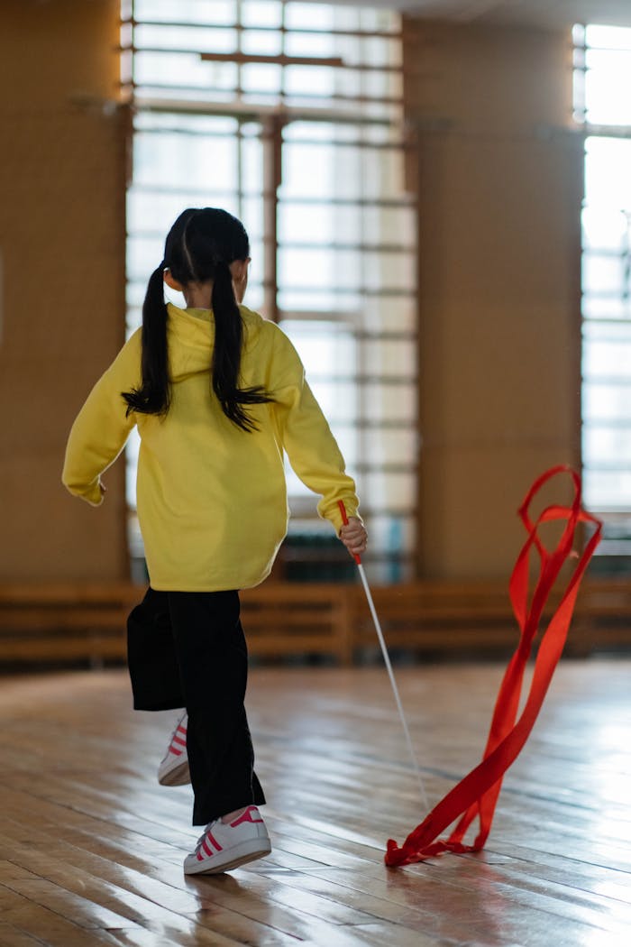 A young girl in a yellow shirt performs gymnastics with a ribbon on a wooden floor indoors.